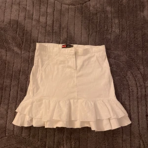 Vit volangkjol med broderad kant - Supersöt vit kortkjol  med dubbla volanger och broderad nederkant. Kjolen har fickor fram och är gjord i bomull, perfekt för en fräsch och somrig look. vet inte vart är ifrån men köpte sc.💕