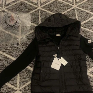 Svart Moncler pufferjacka med huva - Hjälper min kille själva sin snygg svart pufferjacka från Moncler med huva och stickade ärmar. Jackan har dragkedja framtill, två fickor med dragkedja och klassisk Moncler-logga på ärmen. Perfekt för kyliga dagar när du vill ha både stil och värme. Lappar och påse finns tyvärr inte kvar då han har slängt dom men bevis finns på att den är äkta,  vi tar detta seriöst då han inte vill ha den längre så skriv gärna om du verkligen vill ha den. Pris går att diskutera!!!! Skriv för mer info. 