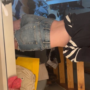 Högmidjade jeansshorts med slitningar - Snygga högmidjade jeansshorts i klassisk blå denim med slitna detaljer och fransig kant. Shortsen har stora bakfickor och knäppning fram med knapp. Perfekta för en avslappnad och trendig look under varma dagar.