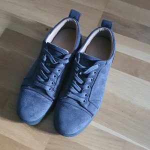 Grå mockasneakers från Christian Louboutin - Snygga sneakers i grå mocka från Christian Louboutin med klassisk röd sula och rund tå. Skorna har snörning och diskreta detaljer, perfekt för dig som gillar stilrena och exklusiva sneakers med en twist.