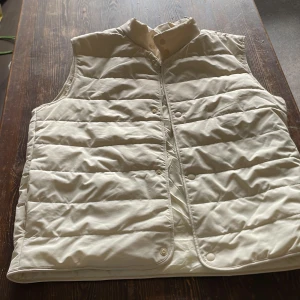 Beige quiltad dunväst från Zara - Snygg beige dunväst från Zara med quiltad design och hög krage. Västen har tryckknappar framtill och är perfekt att slänga över en hoodie eller tröja för extra värme. Enkel och stilren look som funkar till många outfits.