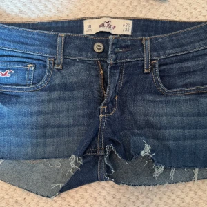 Blå jeansshorts från Hollister - Snygga blå jeansshorts från Hollister med råa, fransiga kanter och klassiska fem fickor. Shortsen har låg midja och cool slitning framtill. Perfekta för sommaren och ger en avslappnad vibe. Broderad Hollister-logga på fickan och dekorativa sömmar bak. Midjemått 40cm