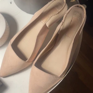 Beige pumps med spetsig tå - Ljus rosa Snygga pumps i beige mocka-look . Skorna har spetsig tå, låg klack och öppen häl med elastiskt band för extra komfort. Perfekta för dig som gillar eleganta skor med en feminin touch.