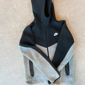 Nike hoodie svart och grå med dragkedja - Nike tech hoodie i svart och ljusgrått med hel dragkedja och huva. Snygg tvåfärgad design med vit Nike-logga på bröstet. Praktiska fickor med dragkedja på sidorna. Perfekt för chill eller träning. Äkta beställd från Nike hemsida. Har bara använt den några gånger ä. 