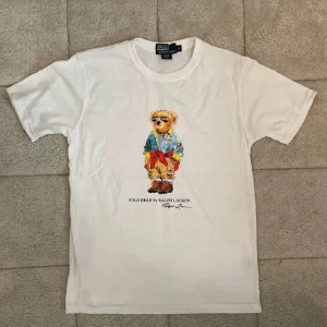 Vit Polo Bear t-shirt från Ralph Lauren - Vit t-shirt från Polo Ralph Lauren med ikoniskt Polo Bear-tryck framtill. Klassisk rund hals och korta ärmar. Tillverkad i mjuk bomull, perfekt för en chill och stilren look. Snygg detalj med färgglad björn som sticker ut.