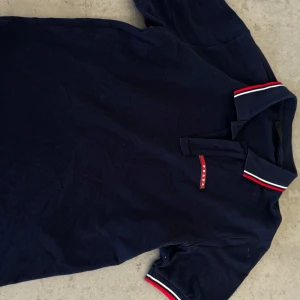 Mörkblå pikétröja Prada - L (sitter som M) - Snygg mörkblå pikétröja från Prada med klassisk krage och knäppning framtill. Kragen och ärmsluten har röda och vita ränder som ger en sportig vibe. Tröjan är tillverkad i mjuk bomull och har diskret Prada-logga på bröstet.