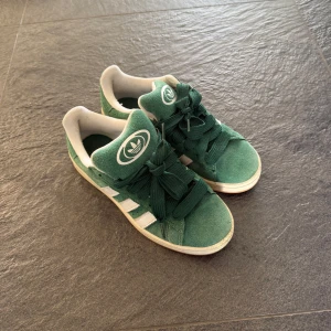 Gröna Adidas Campus - Säljer ett par gröna Adidas Campus sneakers med vita ränder och logga. Smalare gröna och tjockare vita skosnören medföljer. Inga defekter!!