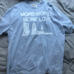 Ljusblå t-shirt med tryck 'More Money More Love' - Säljer en ljusblå t-shirt med vitt tryck på bröstet där det står 'More Money More Love' och en siluett av två personer. T-shirten har en klassisk passform och är kortärmad. Perfekt för en avslappnad stil.