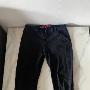 Svarta jeansbyxor från Levi's - Svarta jeansbyxor från Levi's med klassisk femficksdesign och rakt snitt. Byxorna har Levi's-logga på bakfickan och röd logotyp på insidan av linningen. Perfekta för en stilren och enkel look.