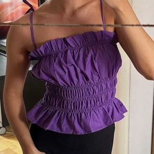 Lila topp - Säljer en lila topp med volangdetaljer och smockad midja. Toppen kan bäras både med och utan axelband och har en luftig, somrig känsla. Perfekt för varma dagar och enkel att styla till olika outfits. Aldrig använd! 