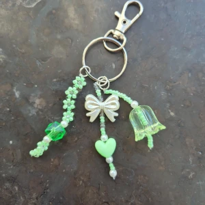 Grön pärlad nyckelring med rosett och hjärta - Unik handgjord nyckelring med gröna och vita pärlor, dekorerad med en söt rosett, ett hjärta och en blomformad pärla. Perfekt accessoar att hänga på väskan eller nycklarna för en personlig touch.