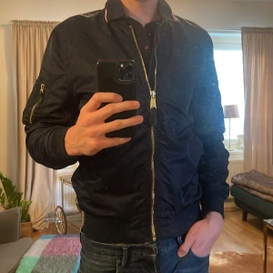 Mörkblå bomberjacka från Alpha Industries - Säljer en klassisk mörkblå bomberjacka från Alpha Industries med guldfärgad dragkedja och fickor på ärmen. Jackan har ribbade muddar vid ärmslut, krage och nederkant. Perfekt för dig som gillar streetwear och enkel stil. 🟢Pris är inte hugget i sten🟢
