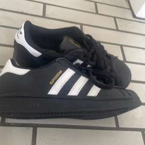 Adidas Superstar sneakers svart/vit - Säljer ett par klassiska Adidas Superstar sneakers i svart med vita ränder och detaljer. Skorna har snörning, platt sula och den ikoniska gummitån. Perfekta för dig som gillar streetstyle och tidlös design.