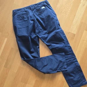 Blå jeans från Gant - Säljer ett par klassiska blå chinos från Gant. Byxorna har normal passform, raka ben och bälteshällor. Perfekta för en stilren och avslappnad look. Fickor bak med knappstängning och diskret logga vid linningen.