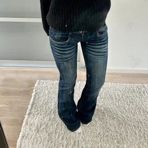 Lågmidjade bootcut jeans - Säljer ett par klassiska blå jeans med låg midja och bootcut passform. Jeansen har snygga slitningar och kontrastsömmar, samt fem fickor och dragkedja med knapp. Perfekta för en avslappnad stil. Gulfen är trasig och går inte att stänga, men ingentingen som syns då dom är så lågmidjade. Säkert lätt att fixa