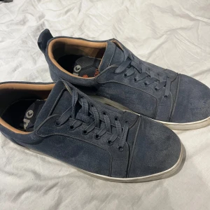 Mörkblå sneakers från Christian Louboutin - Säljer ett par mörkblå sneakers från Christian Louboutin med klassisk röd sula och ovandel i mocka. Skorna har snörning, rund tå och vit platt sula. Insidan är beige och de har diskreta ventilationshål på sidan.
