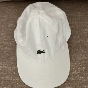 Vit keps från Lacoste - Snygg vit keps från Lacoste med klassisk krokodillogga broderad framtill. Kepsen har böjd skärm och justerbar passform. Tillverkad i bomull och passar perfekt till sommaren.