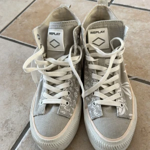 Grå höga sneakers från Replay - Säljer ett par gråa höga sneakers från Replay med vita snören och blommigt mönster på sidorna. Skorna har rund tå, platt sula och tydliga Replay-loggor på plös och insida. Perfekta för dig som gillar stilrena men ändå unika sneakers.