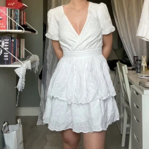 Vit omlottklänning med broderade detaljer - Superfin vit kortklänning med omlottliv och v-ringning. Klänningen har puffiga kortärmar, markerad midja och dubbla volanglager i kjolen. Perfekt till studenten eller sommaren!