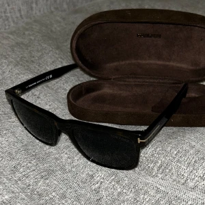 Svarta solglasögon från Tom Ford - Stilrena svarta solglasögon från Tom Ford med fyrkantig form och diskreta gulddetaljer vid gångjärnen. Kommer med originalfodral och putsduk. Perfekta för att lyfta din look under soliga dagar.