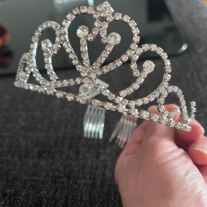 Glittrig tiara med strass - Säljer en vacker tiara i silverfärg dekorerad med gnistrande strass-stenar i ett elegant, hjärtformat mönster. Perfekt accessoar för speciella tillfällen när du vill känna dig extra glam.