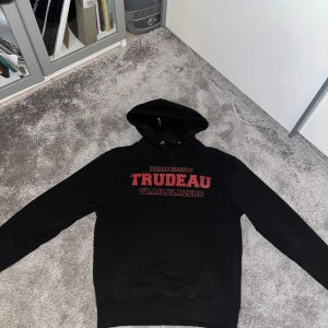 Svart hoodie från Champion med tryck - Säljer en svart hoodie från Champion med rött och vitt tryck på bröstet där det står 'Trudeau Trailblazers'. Klassisk huva och magficka, samt Champion-logga på ärmen. Perfekt för en avslappnad stil.