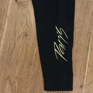 Svarta träningsbyxor från Jordan x PSG - Säljer ett par svarta träningsbyxor från Jordan x PSG med gul Paris-logga på benet och PSG-märke framtill. Byxorna har smal passform och resår i midjan. Perfekta för fotbollsträning eller sportiga outfits.