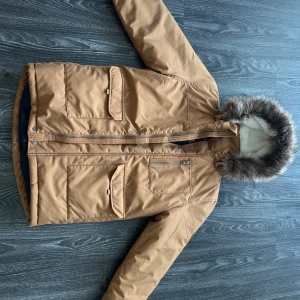 Beige vinterjacka med fuskpäls från Name It - Säljer en beige vinterjacka från Name It med avtagbar huva och fuskpäls. Jackan har flera fickor med lock och dragkedja framtill. Perfekt för kalla dagar och riktigt snygg stil!