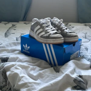 Grå Adidas Campus sneakers med vita ränder - Säljer ett par grå Adidas Campus sneakers med klassiska vita ränder och vit sula. Skorna har snörning och är i mocka med Adidas-logga på hälen. Perfekta för en avslappnad och stilren look.