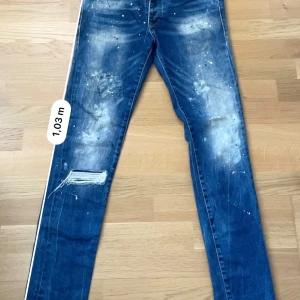 Blå distressed jeans från Dsquared2 - Säljer ett par blå jeans från Dsquared2 med slitna detaljer, färgstänk och en trasig patch på vänster ben. Klassisk femficksmodell med knappgylf och Dsquared2-logga vid midjan. Perfekta för dig som gillar en streetstil.