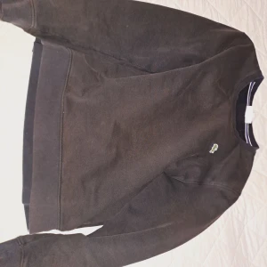 Svart sweatshirt från Lacoste - Klassisk svart sweatshirt från Lacoste med rund halsringning och den ikoniska krokodilloggan broderad på bröstet. Tröjan har långa ärmar och ribbade muddar vid ärmslut och nederkant. Perfekt för en stilren och avslappnad look. Skiftar lite i brunt