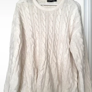 Vit kabelstickad tröja från 157 - Mysig vit kabelstickad tröja från 157 med rund halsringning. Tröjan har ett klassiskt flätmönster och långa ärmar, perfekt för kyliga dagar. Passar dig som gillar en enkel och stilren look.