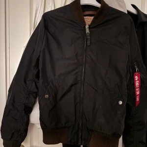 Svart bomberjacka från Alpha Industries - Säljer en klassisk svart bomberjacka från Alpha Industries med orange foder och ribbade muddar. Jackan har dragkedja framtill, två snedställda fickor med tryckknapp och en ikonisk röd tag på ärmen. Perfekt för dig som gillar streetwear.