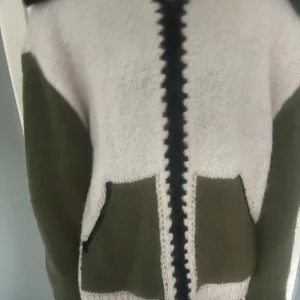 Stickad kofta i vitt och olivgrönt från Zara - Säljer en mysig stickad kofta från Zara i vitt och olivgrönt. Koftan har långa ärmar, två stora fickor framtill och dragkedja. Kragen är bred och stickad i olivgrönt. 