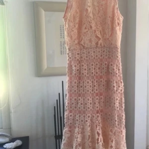 Korallfärgad från True Decadence - Säljer en romantisk persikofärgad, pastell orange klänning från True Decadence med vacker blommig spets och volangdetaljer. Klänningen är ärmlös och har en feminin, figurnära passform med öppen rygg. Perfekt för sommarens speciella tillfällen!