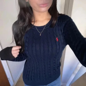 Mörkblå kabelstickad tröja från Ralph Lauren - Säljer en klassisk mörkblå kabelstickad tröja från Ralph Lauren med rund halsringning och röd broderad logga på bröstet. Tröjan har lång ärm och är i mjuk bomull, perfekt för en stilren vardagslook.