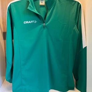 träningströja från Craft - Säljer en grön långärmad träningströja från Craft med vit logga på bröstet och vita detaljer längs axlarna. Tröjan har halv dragkedja framtill och är tillverkad i ett syntetmaterial som passar perfekt för sport och aktivitet. Helt ny. By pris 449