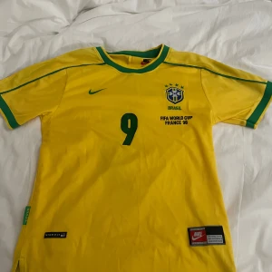 Brasilien fotbollströja Ronaldo 9 FIFA World Cup 1998 - Gul Brasilien fotbollströja från Nike med gröna detaljer, nummer 9 och Ronaldo på ryggen. Officiell FIFA World Cup France 98-design med broderad landslagssköld och Nike-logga. Klassisk retrostil för fans av Ronaldo och Brasilien. Helt ny aldrig använd, storlek S, priset kan diskuteras