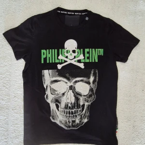 Philipp plein Svart t-shirt  - Cool svart t-shirt från Philipp Plein med stor dödskalle och korslagda ben i vitt framtill. Grön logotyp ovanför motivet. Perfekt för dig som gillar statement-plagg och streetstyle. Storlek M, i mycket fint skick!