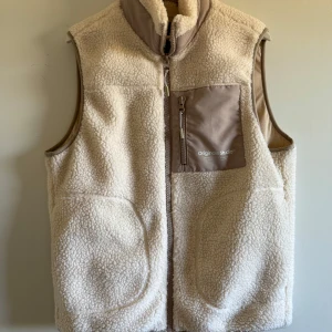 Beige fleeceväst från Jack & Jones - Säljer en beige fleeceväst från Jack & Jones Originals med dragkedja och en bröstficka i kontrasterande tyg. Västen har hög krage och är perfekt för lager-på-lager-stil. Enkel och stilren design med mjukt material. Knappt använd. Storlek s