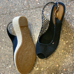 Marinblå kilklackar från Tommy Hilfiger - Säljer ett par marinblå sandaler från Tommy Hilfiger med öppen tå och kilklack i repimitation. Skorna har ett snyggt mönstrat innerfoder och en liten logga på sidan. Perfekta för sommaren!