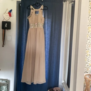 Beige långklänning med paljettdetaljer - Säljer en elegant beige långklänning med glittrande paljetter runt midjan och halsringningen. Klänningen har breda axelband och ett vackert fall i kjolen. Perfekt för speciella tillfällen när du vill sticka ut.