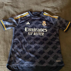 Real Madrid bortatröja 2023/24 från Adidas - Säljer en mörkblå Real Madrid bortatröja från Adidas med gula detaljer och mönstrad framsida. Tröjan har korta ärmar, klubbmärke och sponsortryck på bröstet samt 'REAL MADRID' i nacken. Perfekt för fotbollsfans! Pris kan diskuteras.