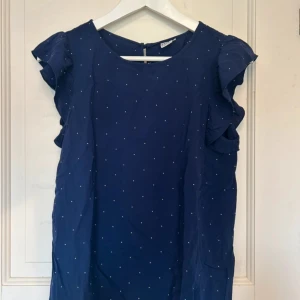 Mörkblå topp med volangärmar - Söt mörkblå topp med små vita prickar och volangdetaljer vid ärmarna. Rund halsringning och lätt, luftig passform. Perfekt till sommaren