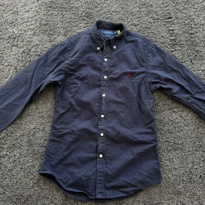 skjorta från Polo Ralph Lauren - Säljer en mörkblå skjorta från Polo Ralph Lauren med slim fit-passform. Skjortan har långa ärmar, knappar framtill och klassisk krage med knapp. Liten röd broderad logga på bröstet. Perfekt för en stilren look. Aldrig använd och köpt på room 1006 på Emporia.