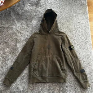 Olivgrön hoodie från Stone Island x Supreme - Säljer en olivgrön hoodie från Stone Island och Supreme med broderad logga på bröstet och ärmen. Stor huva, känguruficka och Supreme-tryck på ryggen. Perfekt för dig som gillar streetwear och exklusiva samarbeten.