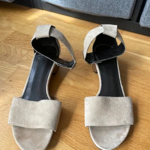 Beige sandaler med klack från Vagabond - Snygga beige sandaler från Vagabond med bred rem över tårna och justerbart spänne vid ankeln. Skorna har en låg klack i trälook och är tillverkade i mocka för en stilren och modern känsla.