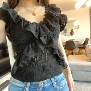 Svart volang topp med spetsdetaljer - Supersnygg svart topp med volanger och spetsdetaljer. Toppen är ärmlös och har en djup v-ringning med dekorativa volanger framtill. Materialet är stickat och spets, vilket ger en romantisk känsla. Perfekt att styla med jeans eller kjol.