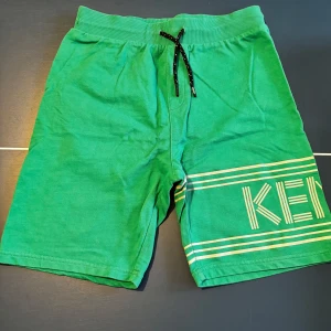 Gröna shorts från Kenzo Kids - Snygga gröna shorts från Kenzo Kids med resår och snörning i midjan. Stora vita Kenzo-loggor med ränder på ena benet ger en cool look. Aldrig använda, Har dock skrivit initialer på etiketten insidan.