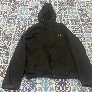 Svart vindjacka från Fred Perry - Svart vindjacka med huva från Fred Perry. Jackan har dragkedja och knappar framtill, två fickor med dragkedja och guldigt Fred Perry-logga på bröstet. Justerbar dragsko i huvan och elastisk nederkant.